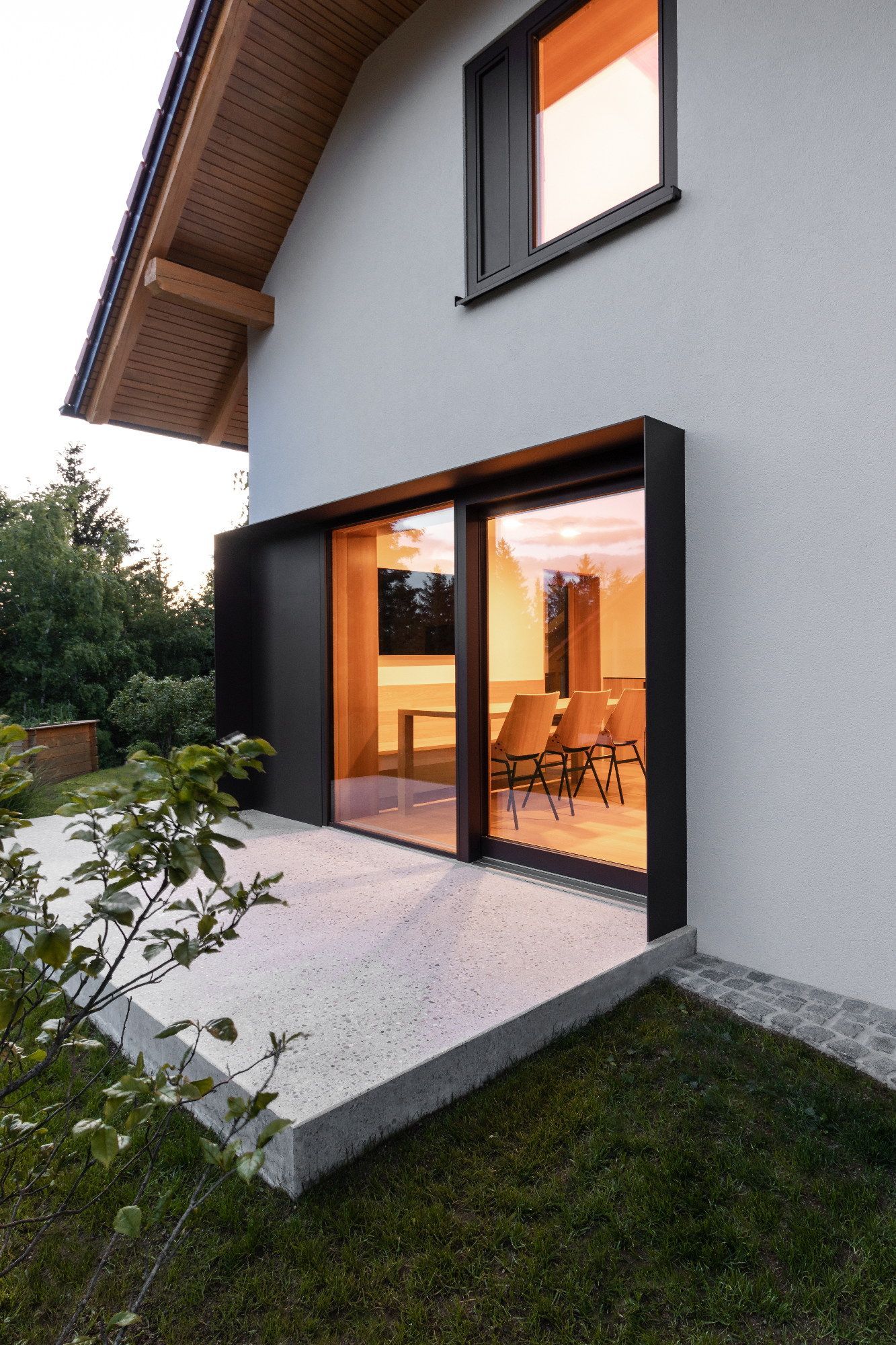 Esterno di una casa moderna con ampie porte scorrevoli in vetro che si aprono su una zona pranzo illuminata e un piccolo prato.