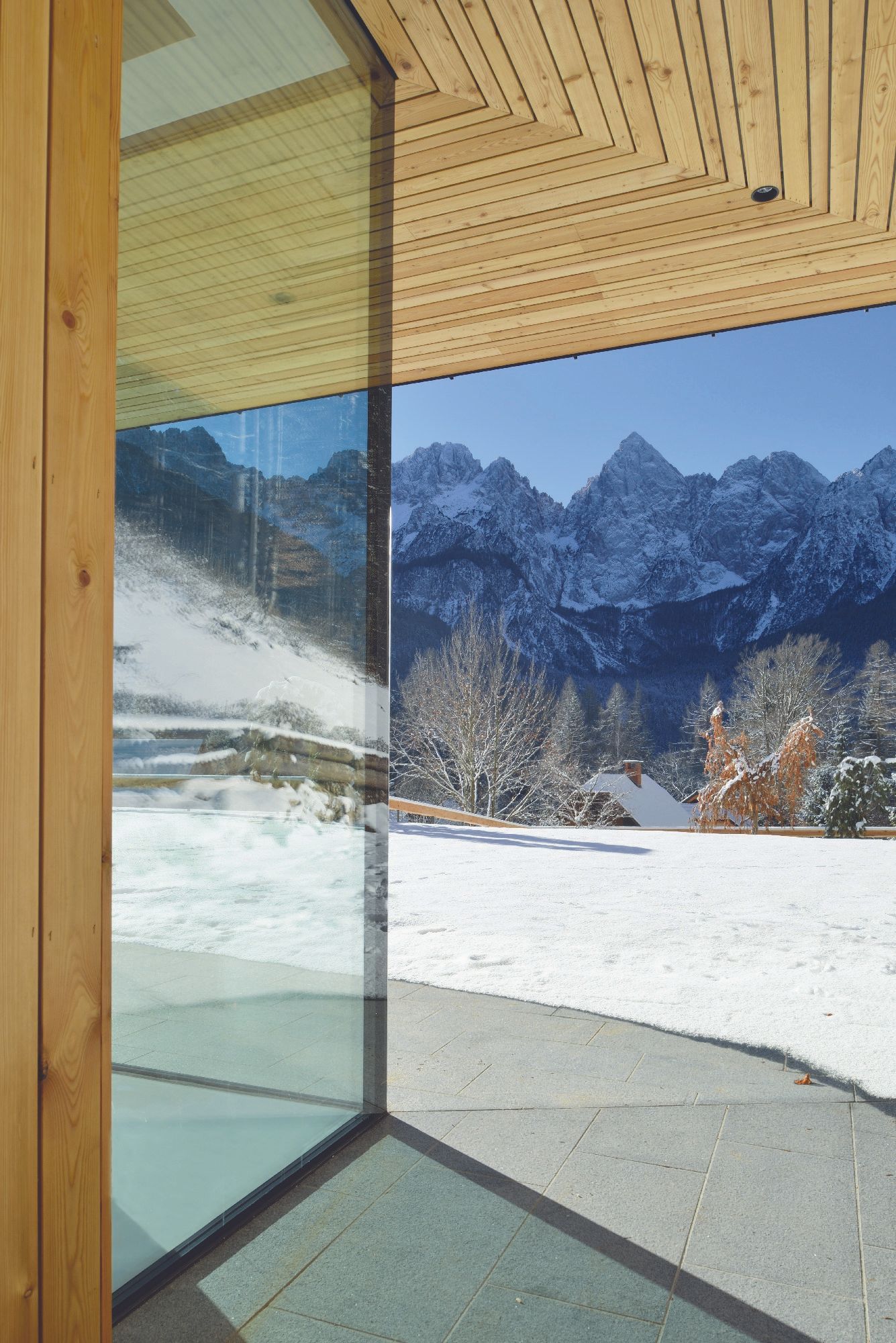 Vista di montagne innevate incorniciata da ampie pareti di vetro e un soffitto in legno da un interno moderno