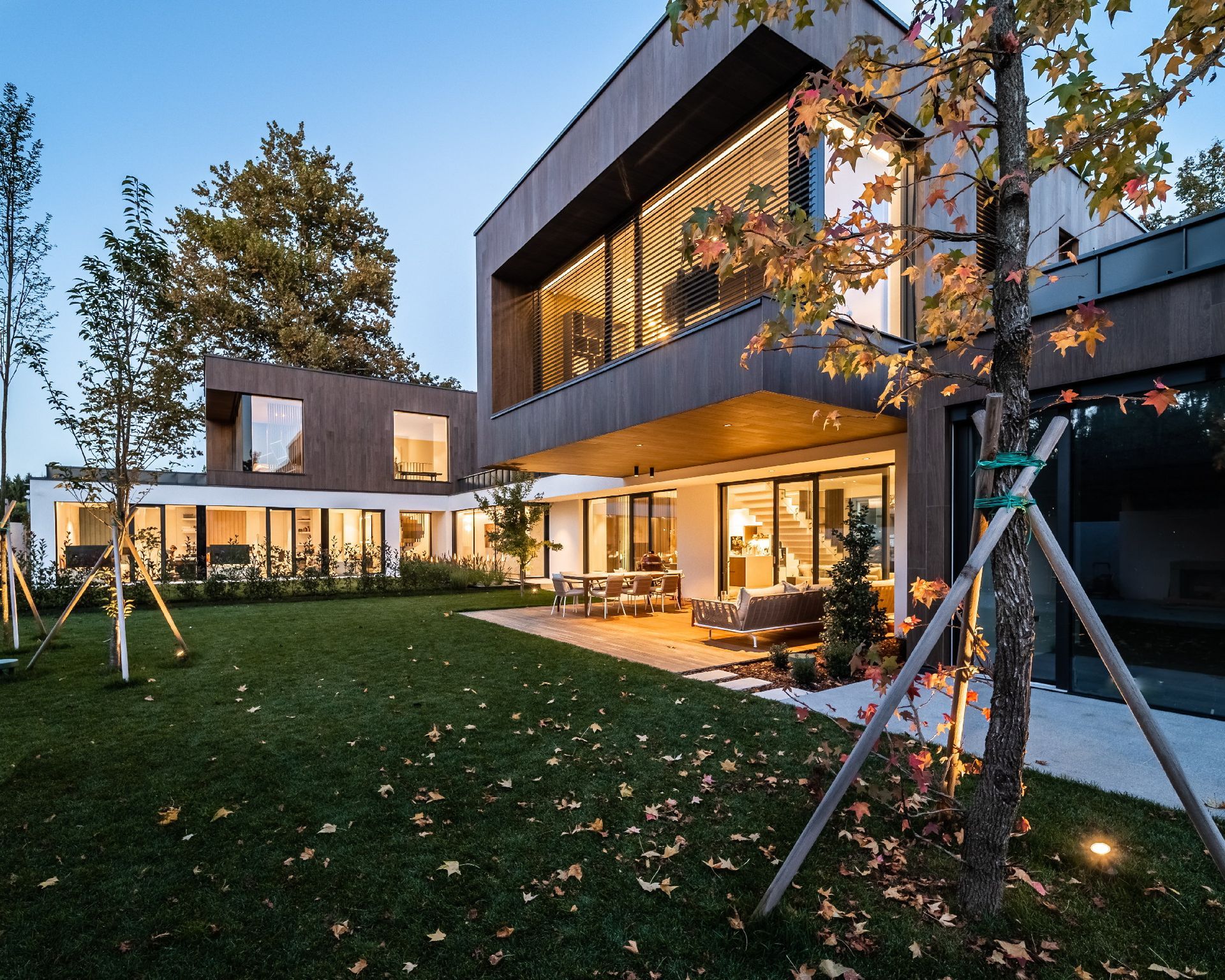 Casa moderna a due piani con ampie finestre e patio illuminato, vista da un giardino erboso al crepuscolo.