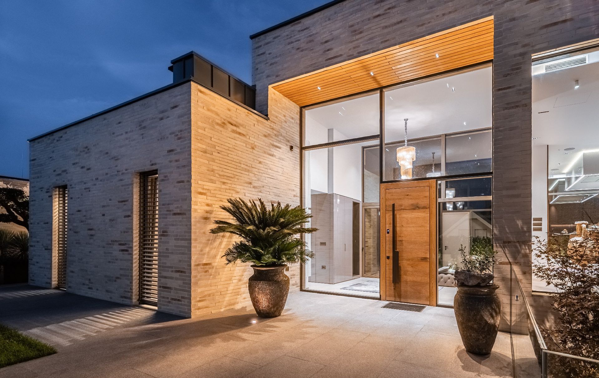 Esterno di una casa moderna al crepuscolo con ingresso in vetro, illuminazione calda e piante in vaso vicino alla porta d'ingresso.