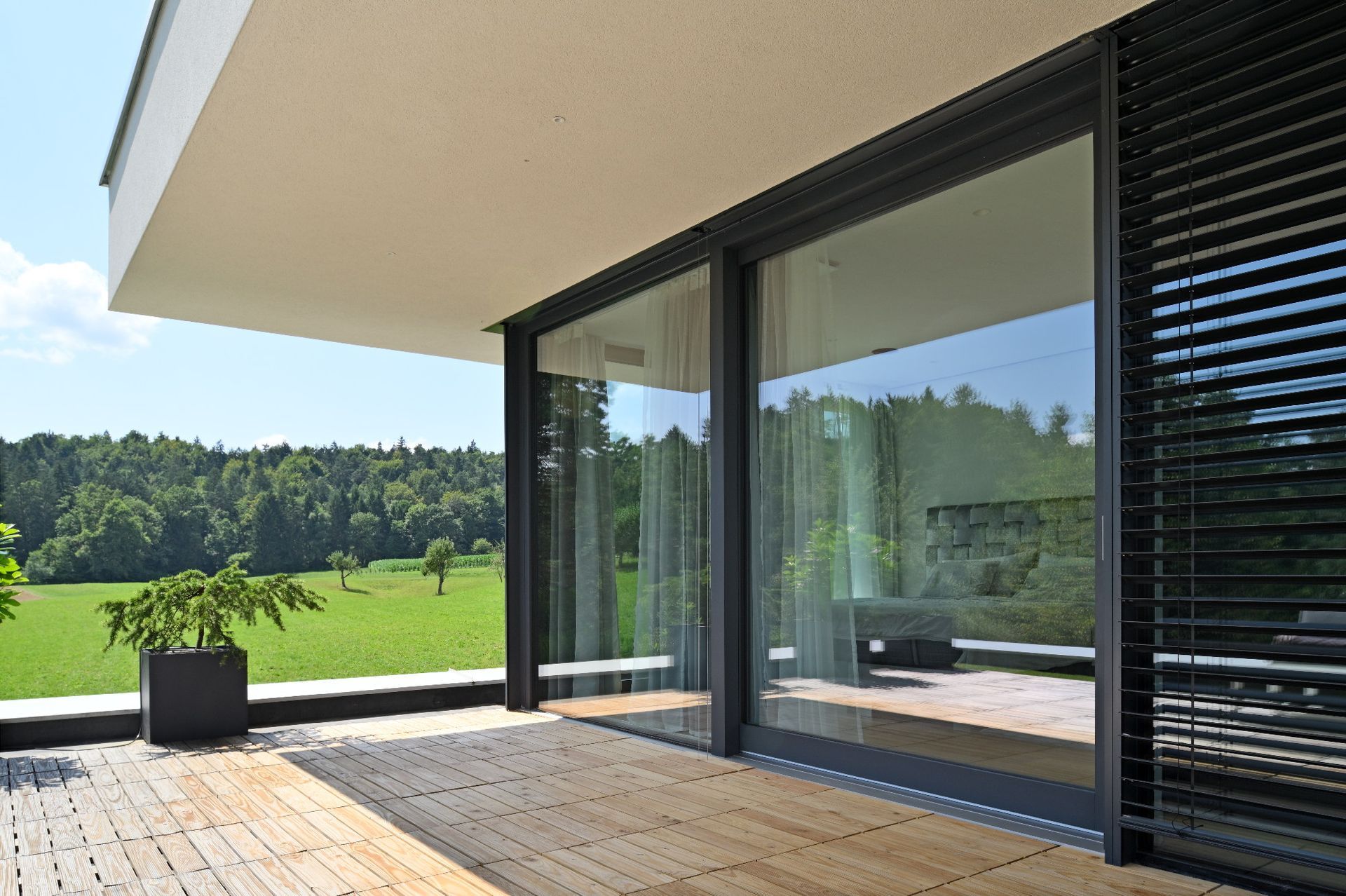 Patio moderno con porte a vetri, pavimentazione in legno e un paesaggio verde e soleggiato sullo sfondo.