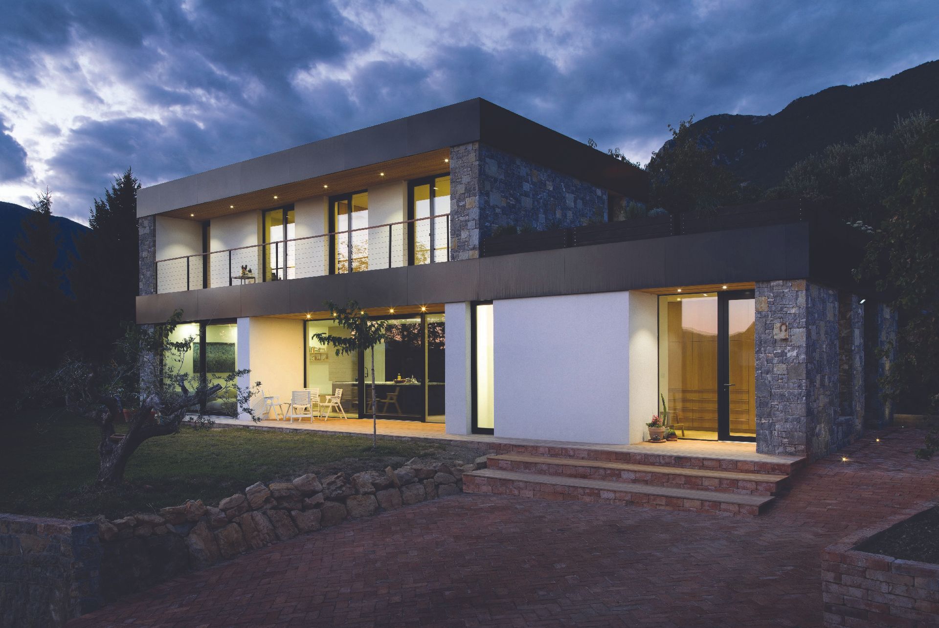 Casa moderna a due piani con ampie finestre che brillano al crepuscolo, incastonata sullo sfondo di una collina scura.
