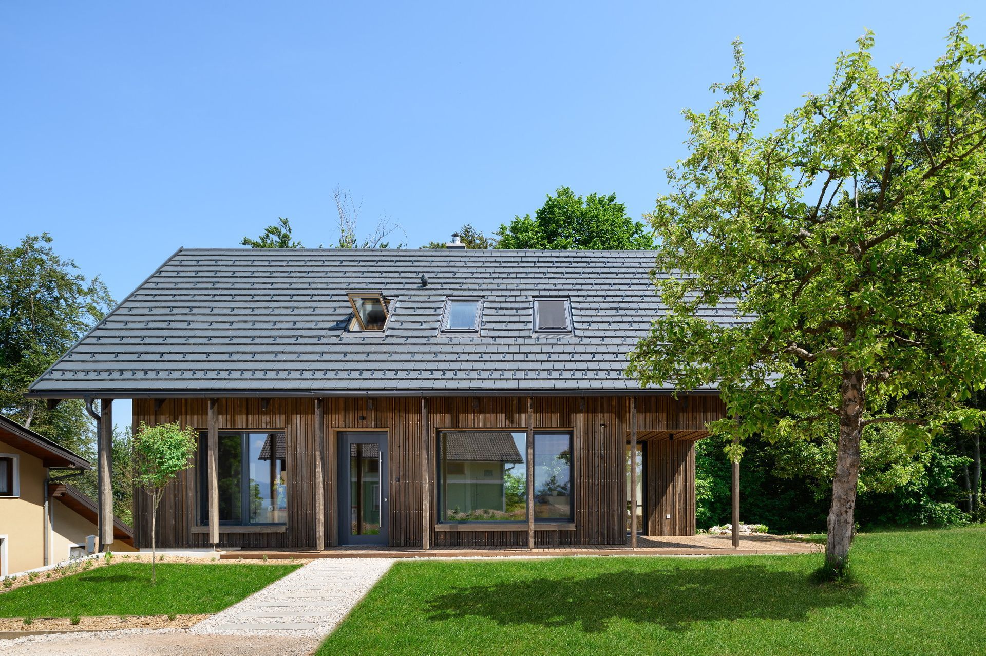 Casa moderna in legno con tetto grigio, grandi finestre e un prato verde sotto un cielo azzurro limpido.