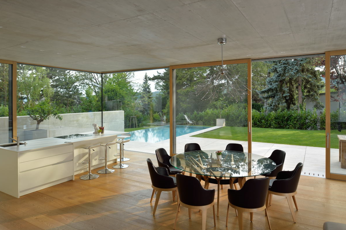 Moderna sala da pranzo con pareti in vetro, tavolo rotondo, sedie nere e vista sulla piscina e sul prato verde.
