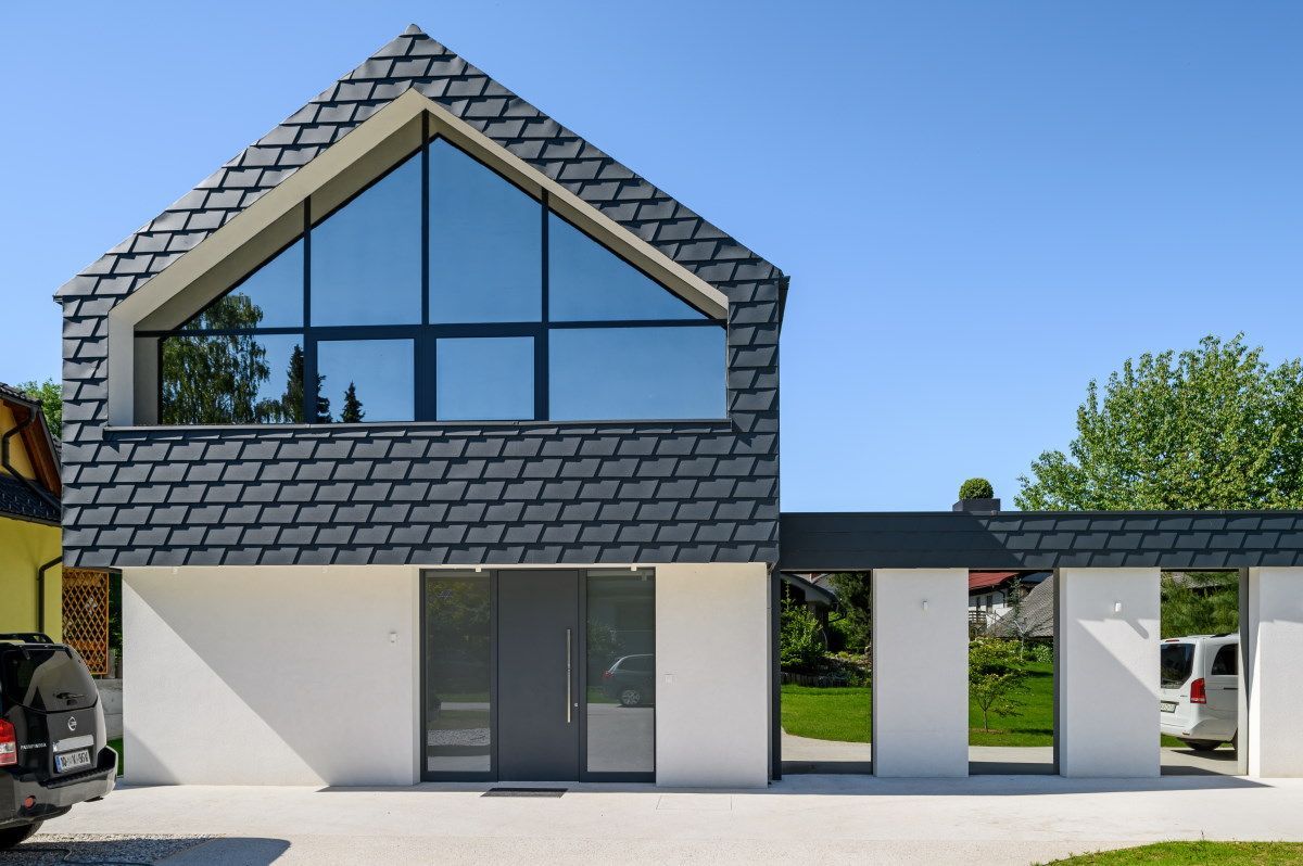 Facciata di una casa moderna con tetto in tegole grigie, grande finestra triangolare e posto auto coperto in una giornata di sole.