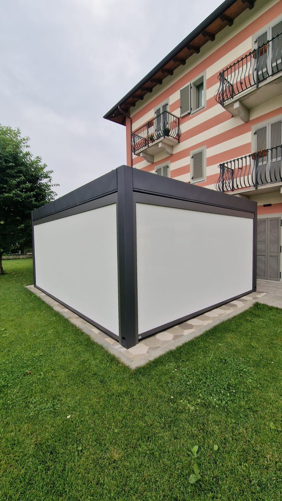 Gazebo bianco da esterno con finiture grigio scuro su un prato accanto a un edificio a strisce.