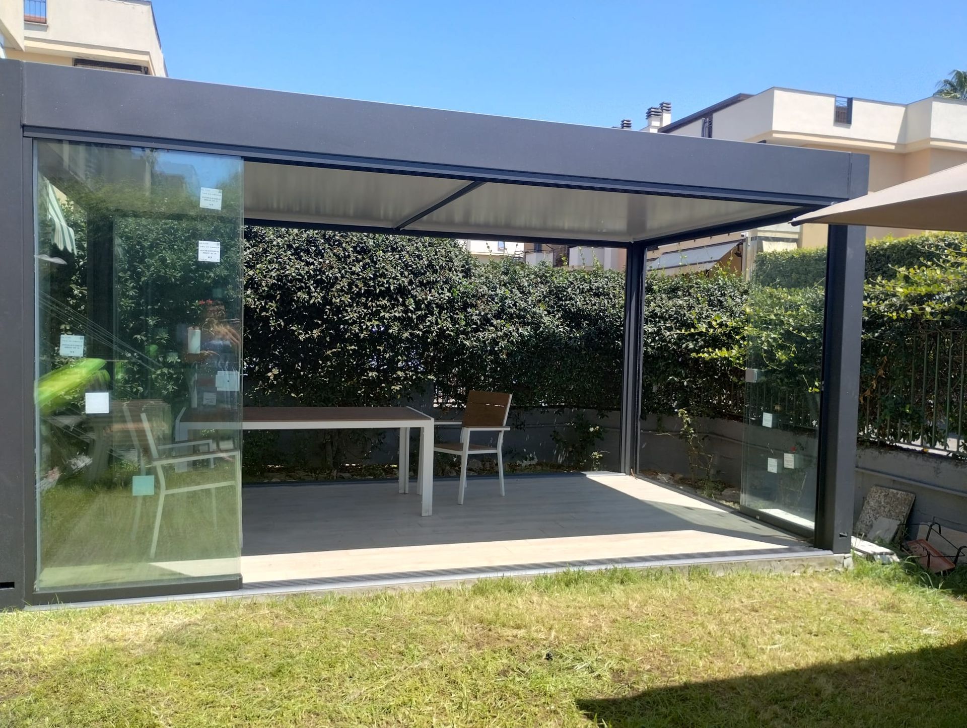 Patio coperto con pareti in vetro, tavolo e sedia bianchi e prato verde antistante.
