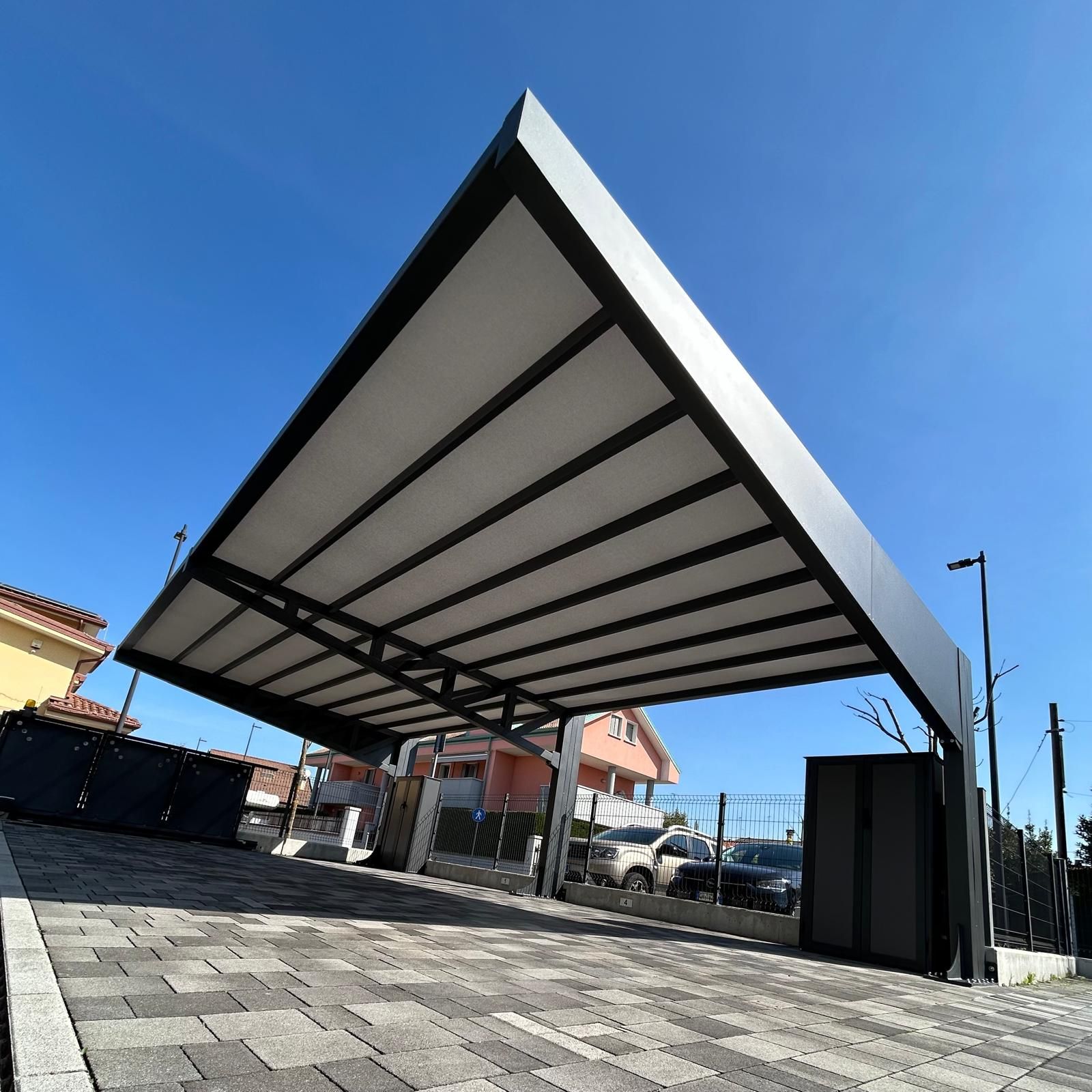 Posto auto coperto moderno con tettoia bianca e supporti neri su un piazzale pavimentato sotto un cielo azzurro.
