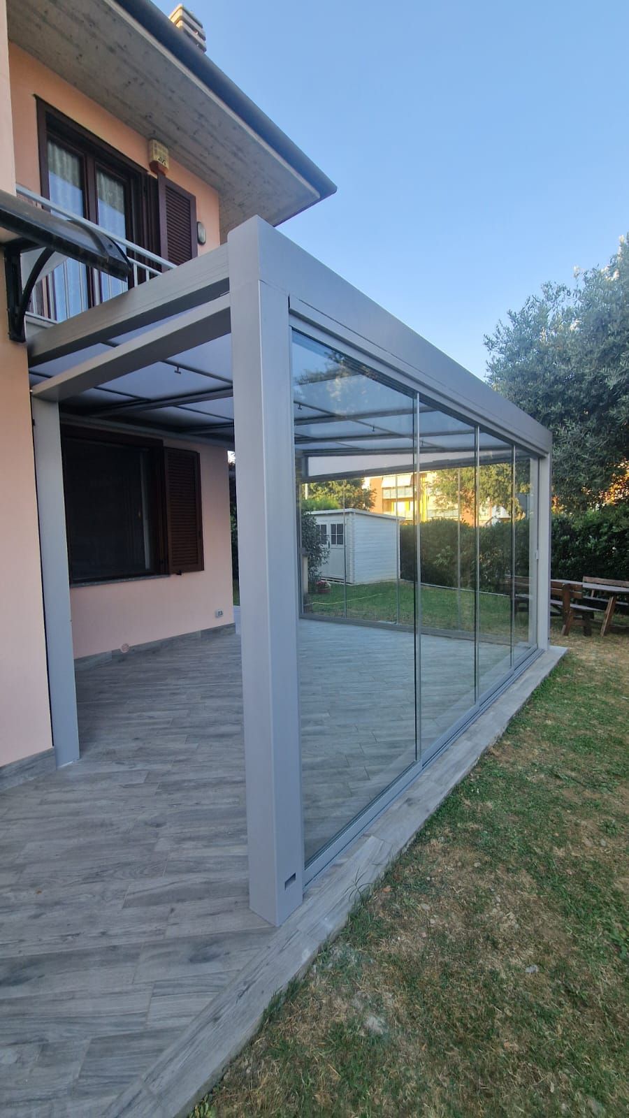 Struttura di pergola in metallo grigio accanto al patio di una casa, con pannelli in vetro e un prato.