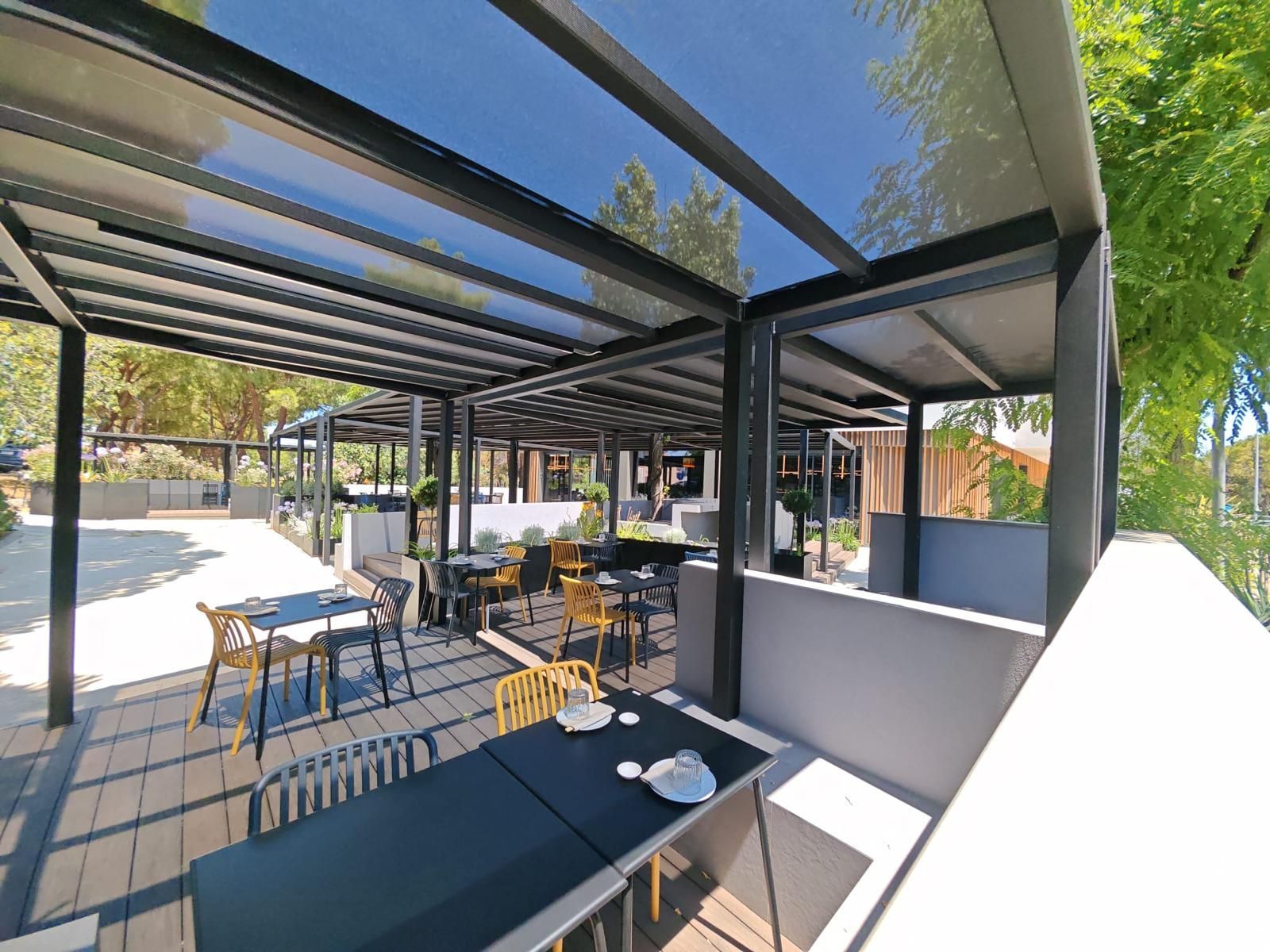 Zona pranzo all'aperto sotto un pergolato nero con tavoli, sedie e luce del sole che proietta ombre a strisce