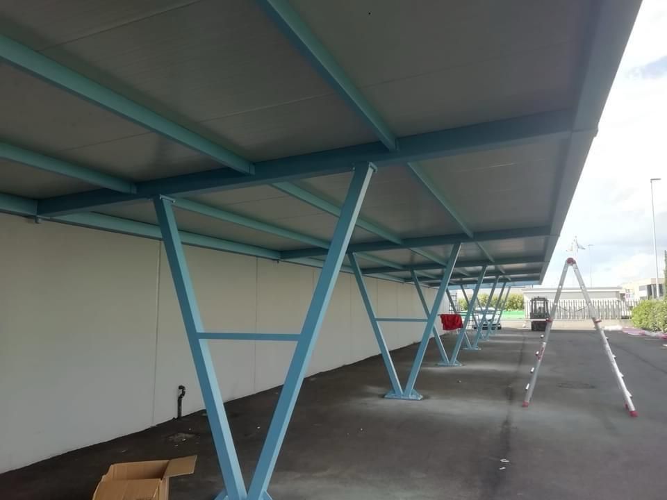 Area di parcheggio coperta con supporti metallici blu e un muro bianco lungo un marciapiede pavimentato.