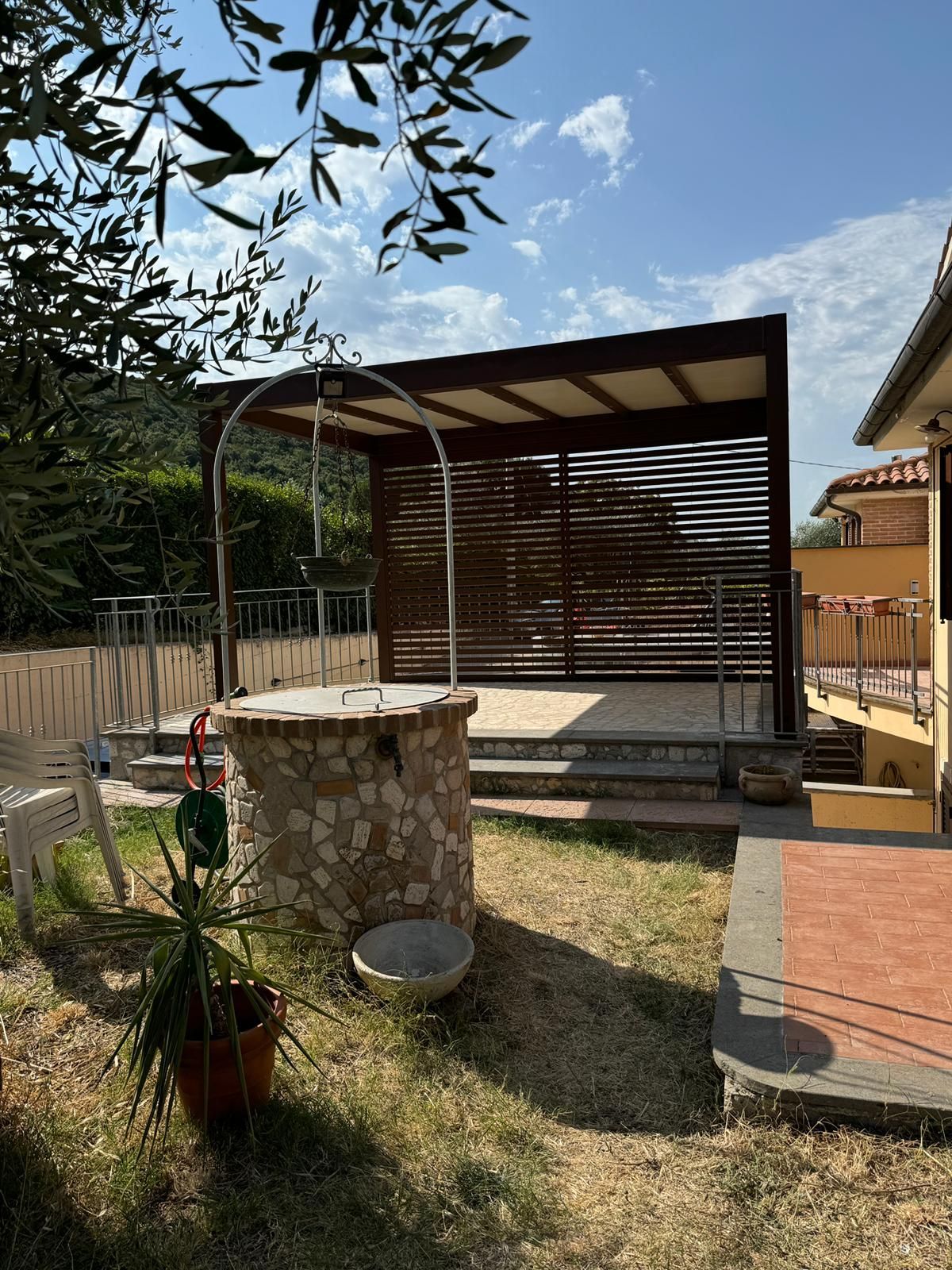 Patio sul retro con pozzo in pietra, pergolato e piante in vaso sotto un cielo soleggiato