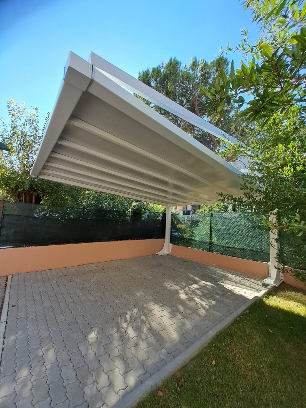 Pergola bianca moderna sopra un patio pavimentato, accanto a prato, alberi e una recinzione verde.
