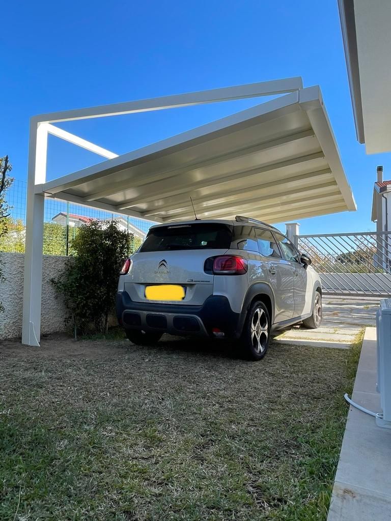 Un SUV bianco è parcheggiato sotto una moderna tettoia in un vialetto soleggiato con ghiaia ed erba.