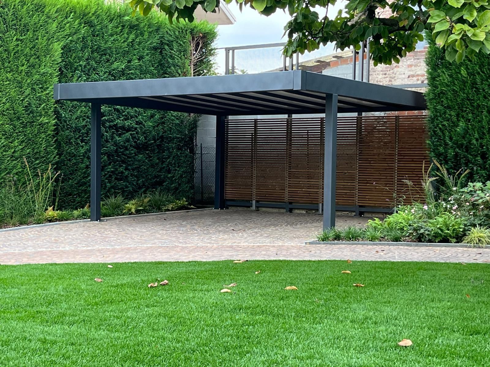 Moderno carport nero con tetto a doghe, accanto a una recinzione in legno e siepi verdi in un giardino.