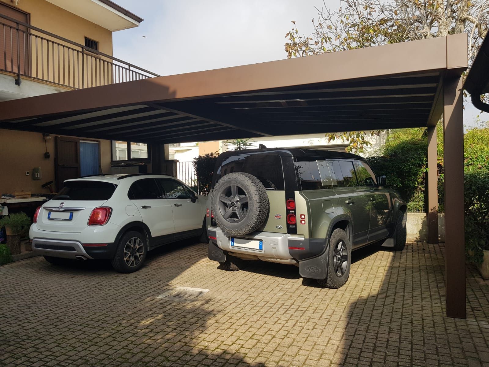Due SUV parcheggiati sotto una tettoia accanto a una casa, uno bianco e uno verde scuro.