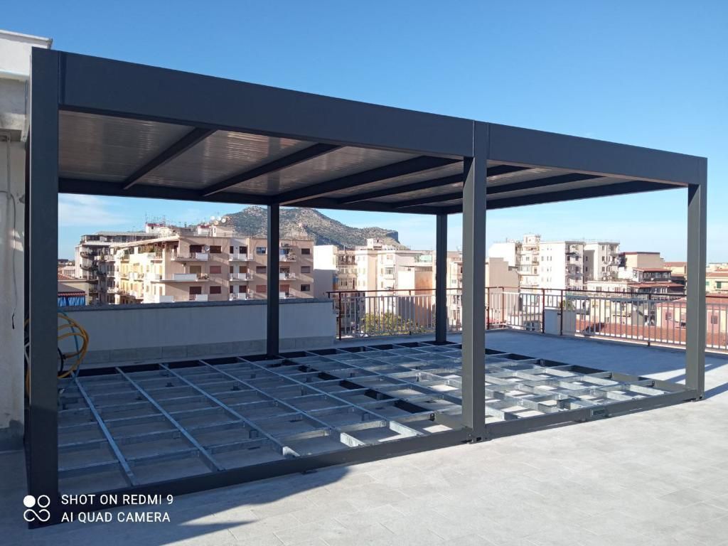 Terrazza sul tetto in costruzione con struttura metallica, pavimento aperto e edifici cittadini sullo sfondo.