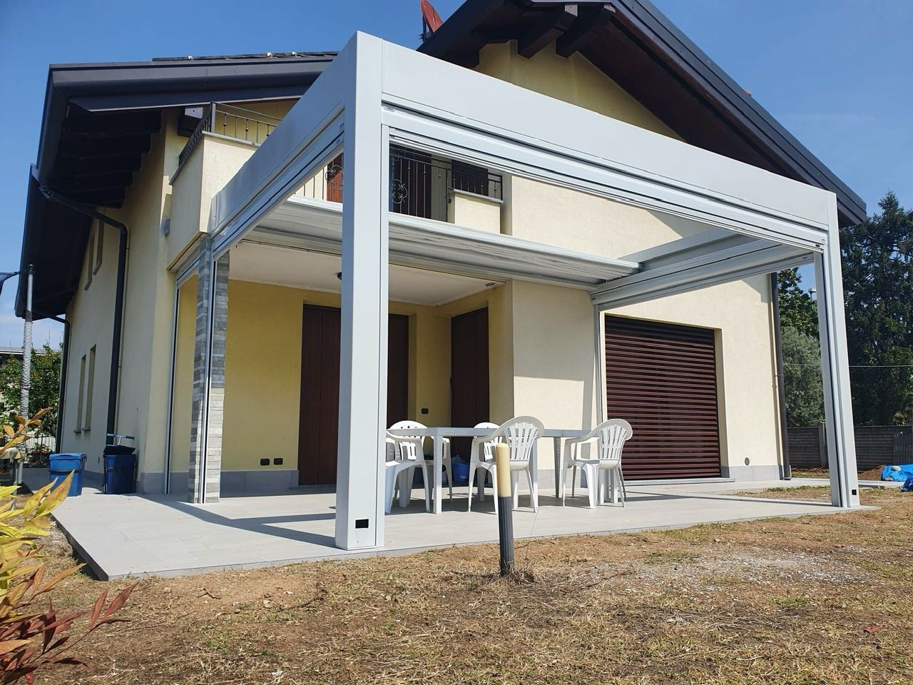 Casa moderna con pergolato bianco sul patio, sedie da esterno e porta del garage marrone sotto un cielo soleggiato