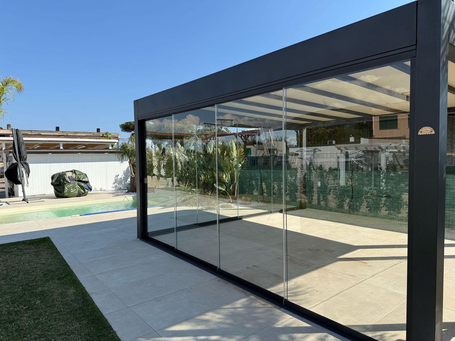 Moderno patio sul retro con veranda vetrata e prato a bordo piscina sotto un cielo limpido.