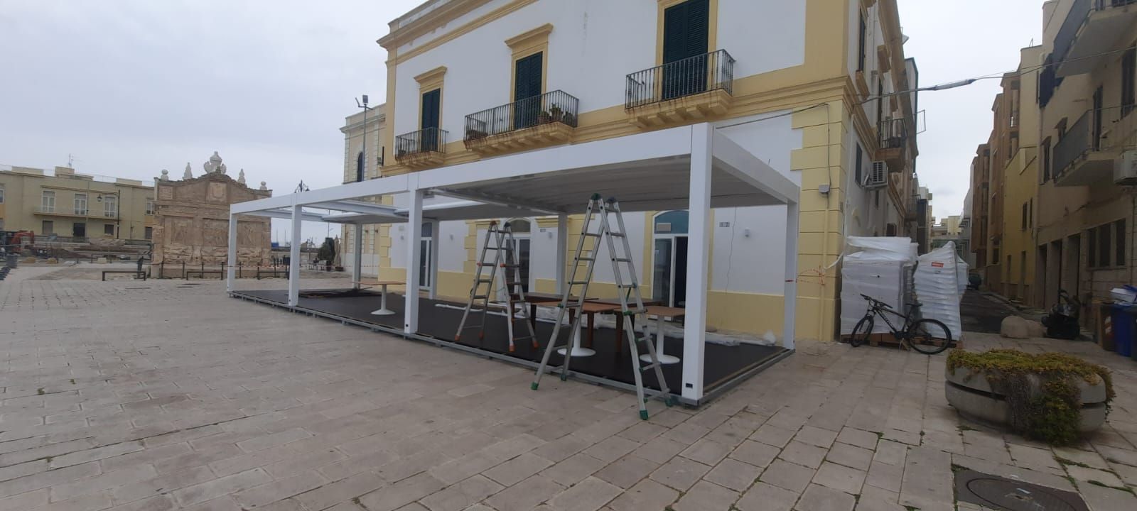 Allestimento di un caffè all'aperto con tendone bianco, tavoli e scalette accanto a un edificio in pietra nella piazza.
