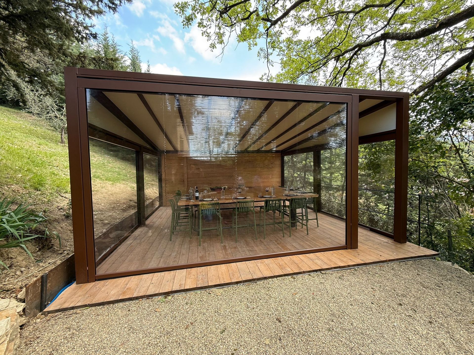 Moderno patio esterno con pareti in vetro, tetto in legno e tavolo da pranzo immerso tra gli alberi.