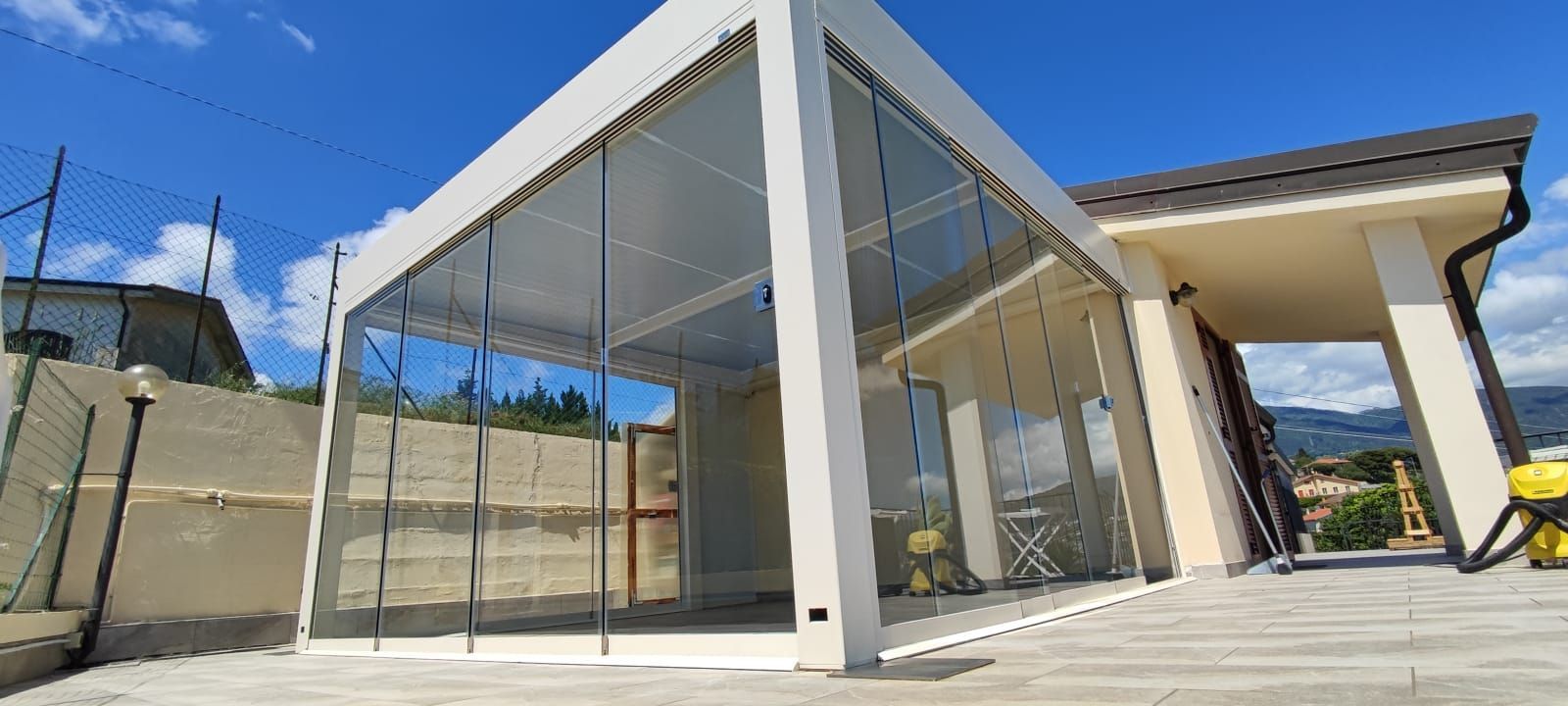 Padiglione moderno in vetro con travi bianche e patio in pietra sotto un cielo azzurro.