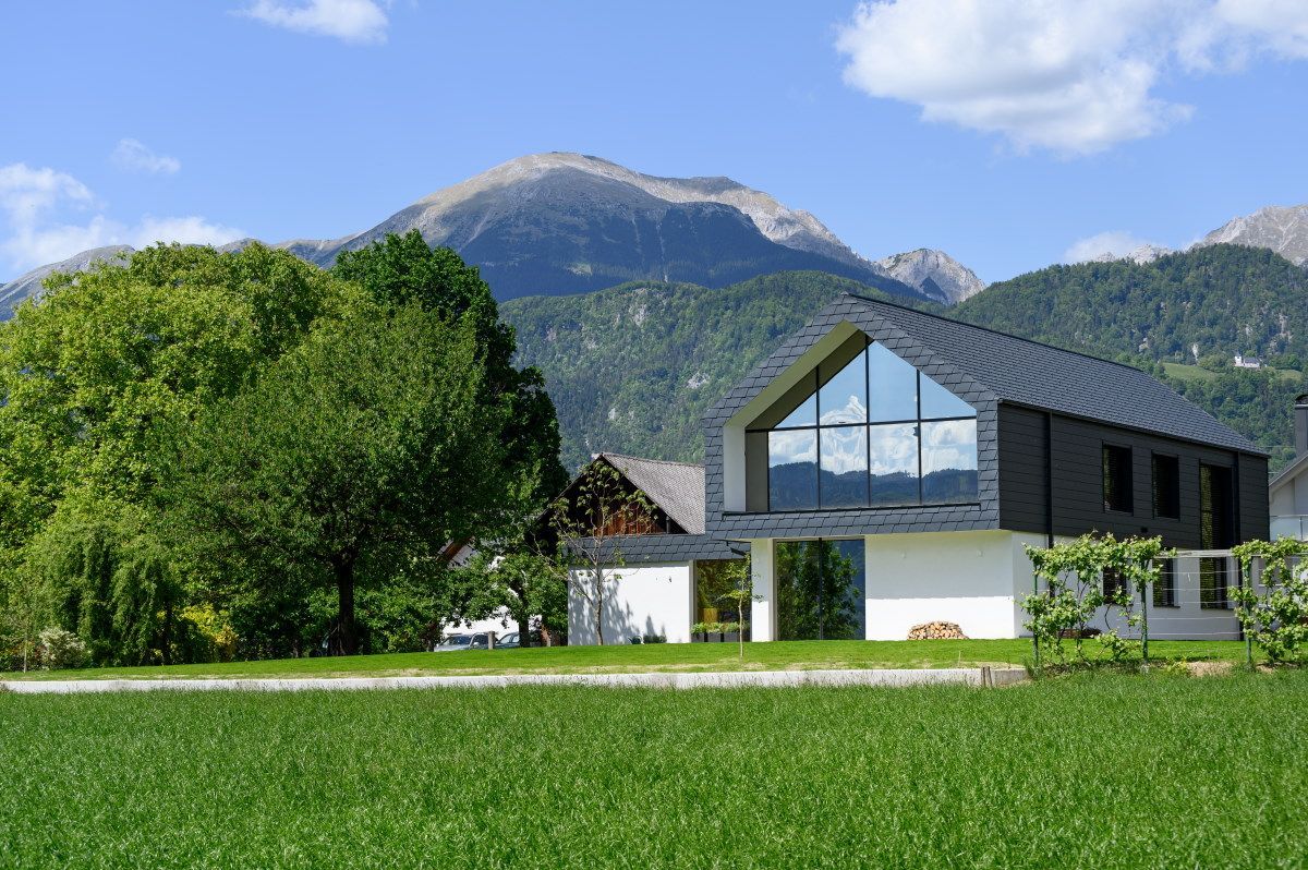 Casa moderna con ampie finestre affacciata su un campo verde, con montagne e cielo azzurro sullo sfondo.