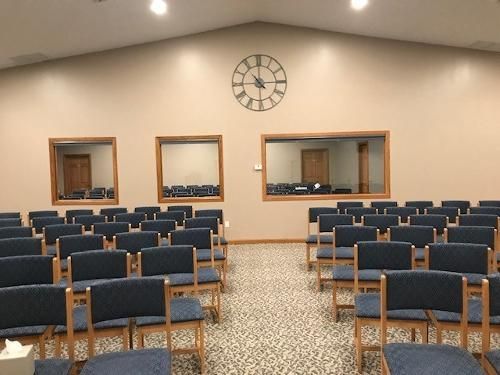 Rows of blue chairs in a beige-walled room with a clock and two windows overlooking another room.