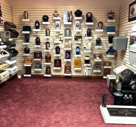Funeral home display of cremation urns on shelves, with pink carpet and memorial items.