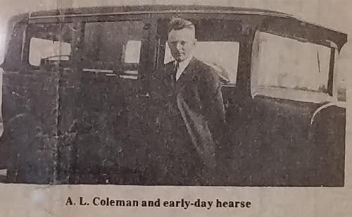 A man in a suit stands by an early-day hearse. Black and white photo.