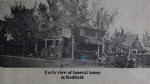 Early view of a funeral home in Redfield; includes a vintage car and surrounding trees.