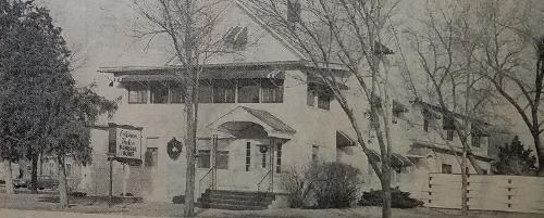 Black and white photo of a two-story building with a sign and trees in front.