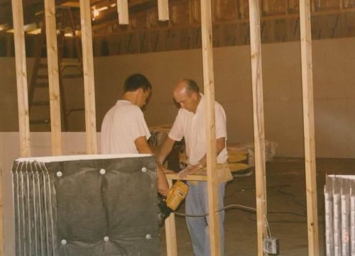 Two men working with wood and a power saw inside a building under construction.