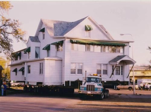 White house on a truck, being moved down a street. Green awnings over the windows.