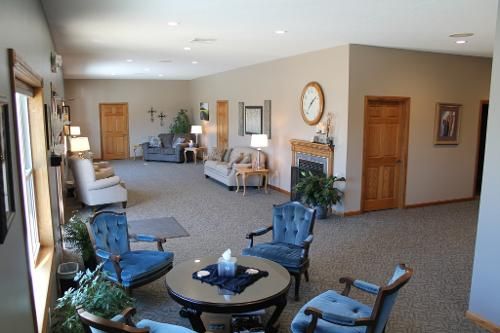 Spacious waiting area with couches, chairs, fireplace, and doors. Blue chairs surround a table. Beige walls, carpet.