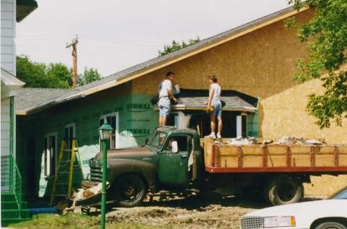 Two people on a truck roof, working on a house with plywood and windows. Green truck parked.