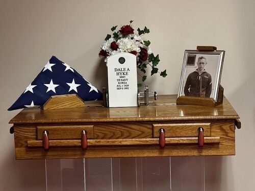Wooden memorial shelf with American flag, photo, flowers, and military tag.