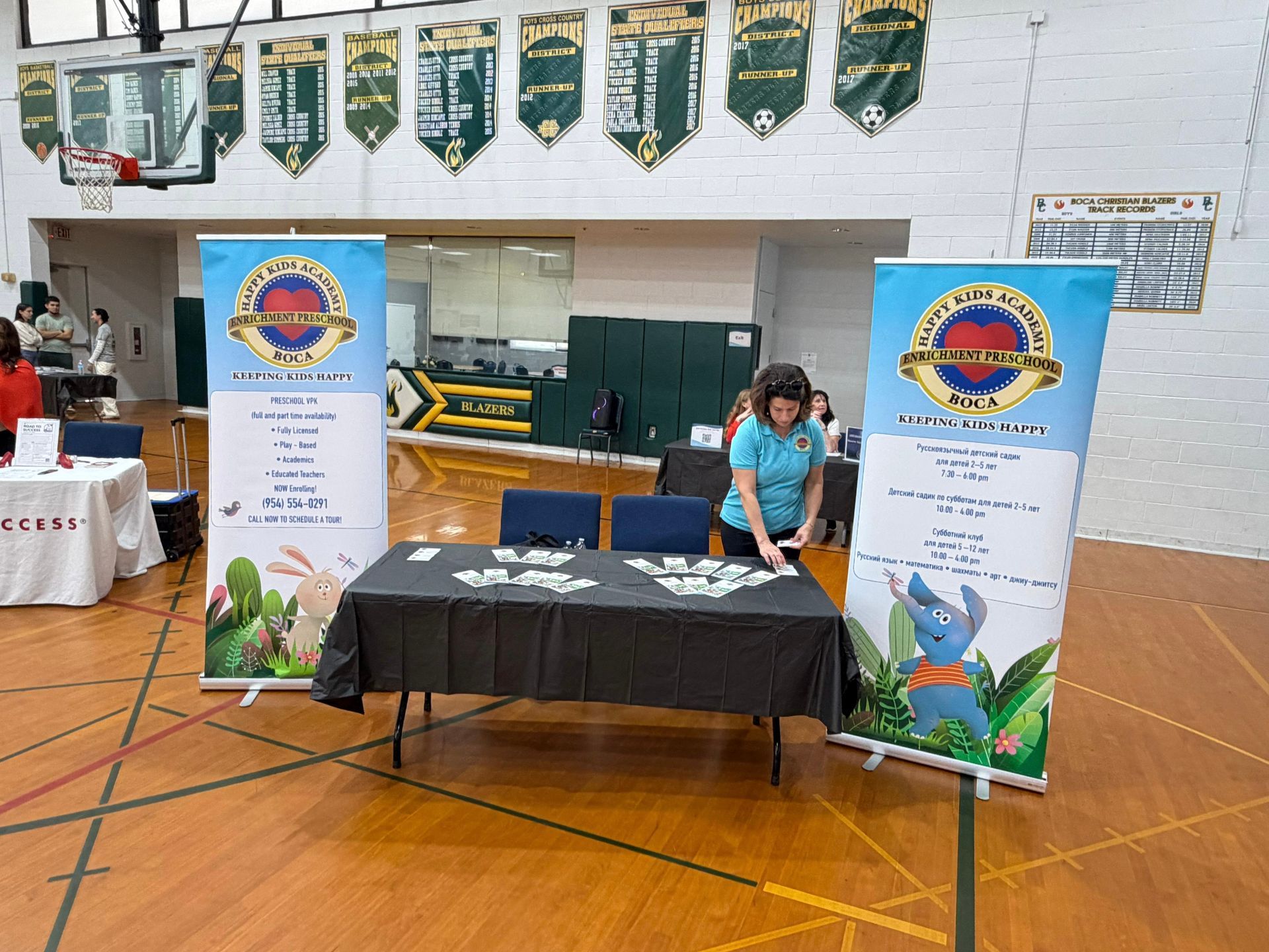 School gym booth with banners and a table, where a masked woman arranges materials.