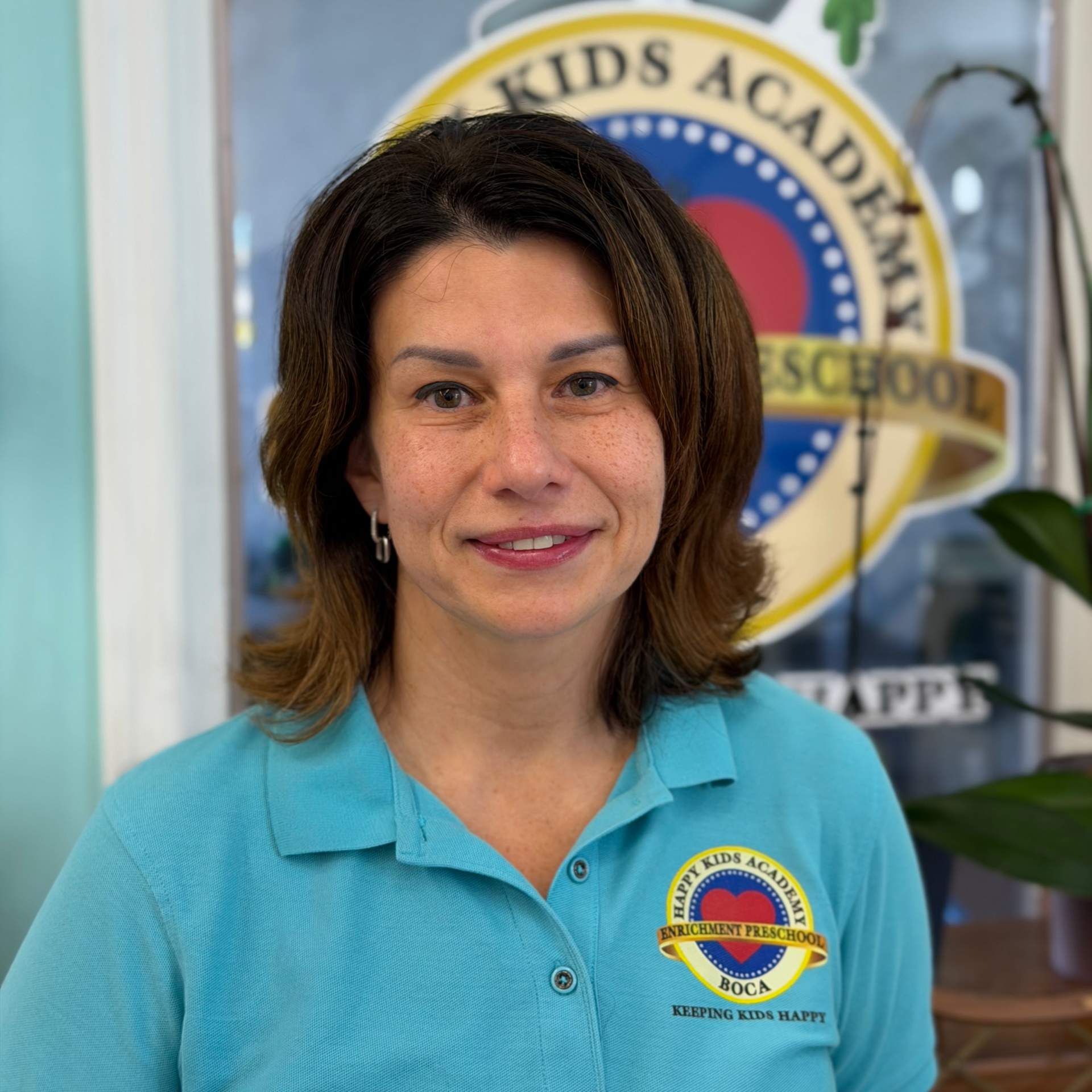 Woman in a turquoise polo shirt standing indoors in front of a school emblem.