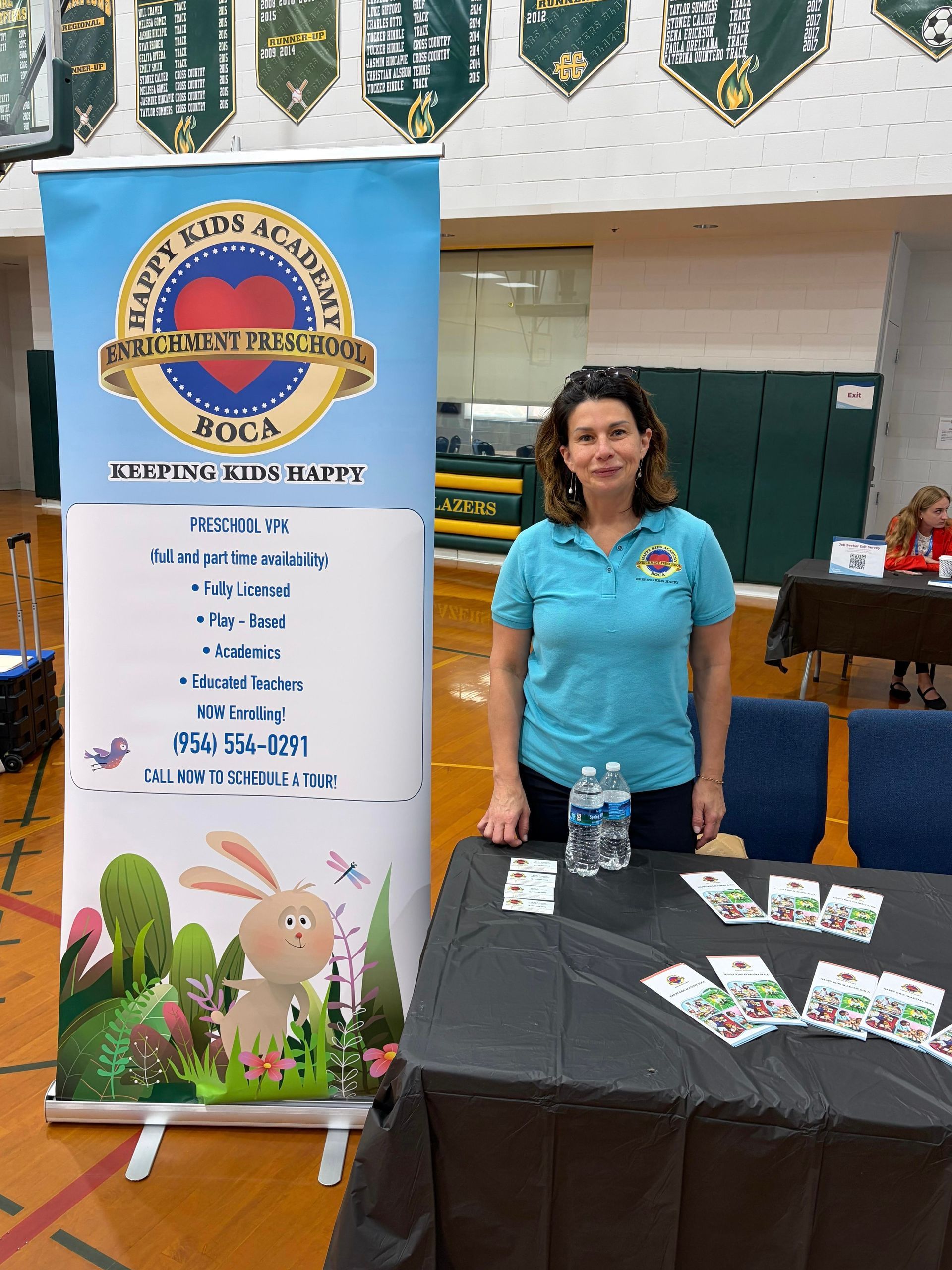 Woman stands beside a promotional banner and table with flyers in a gym-like hall.