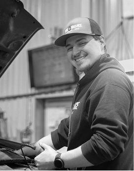 A smiling mechanic wearing a cap and hoodie works under the open hood of a car in a workshop. | Performance Automotive Repair