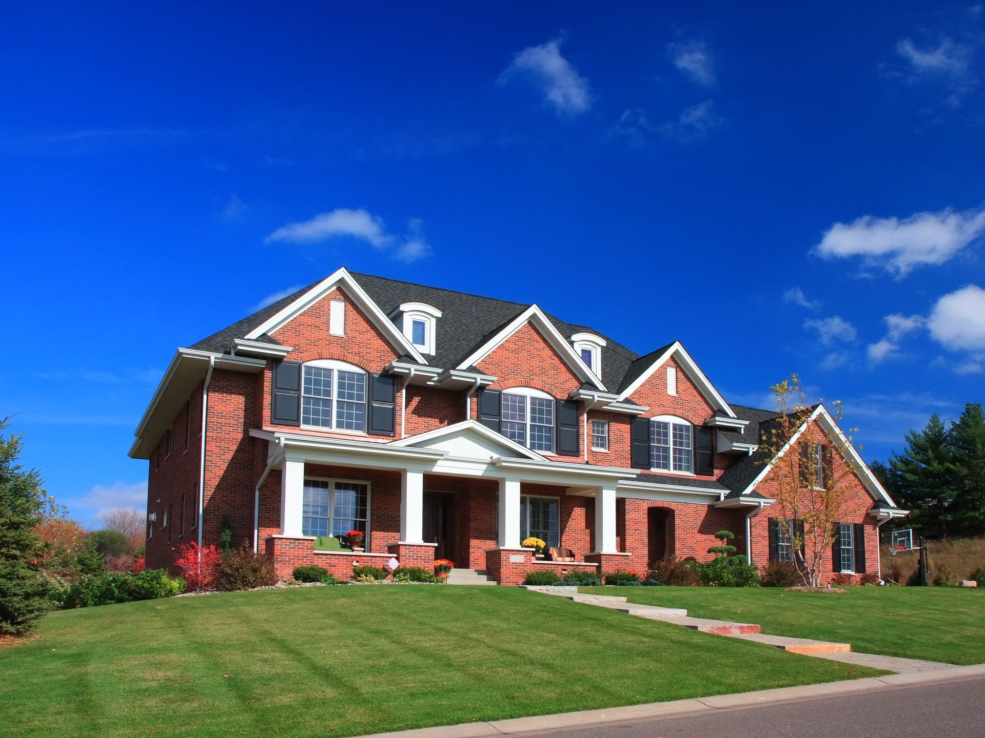 A large, two-story red brick suburban house with a white front porch, black shutters, and a lawn under a bright blue sky.