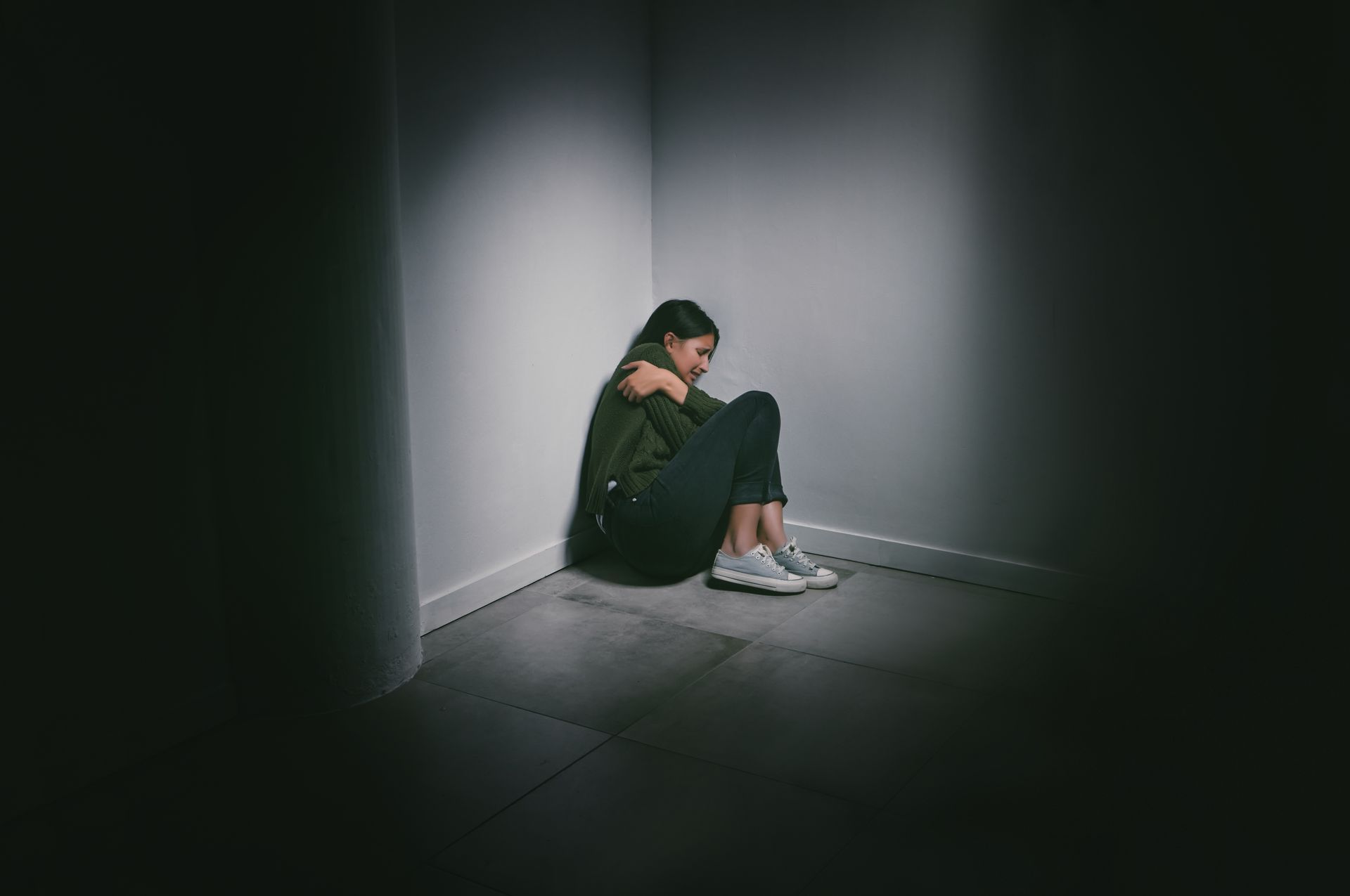 A woman is sitting on the floor in a dark room, hugging her knees.