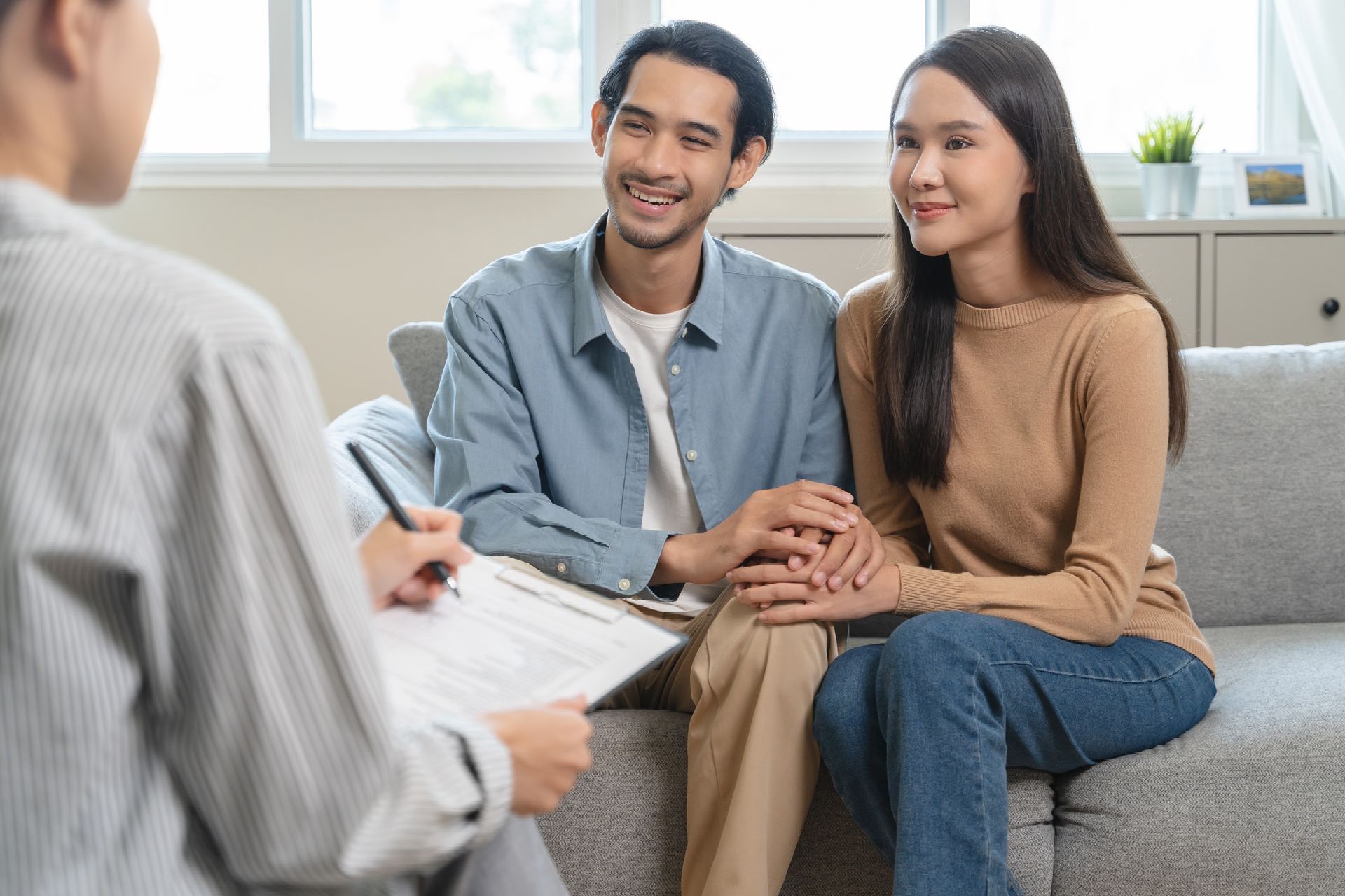 A man is sitting on a couch talking to another man while holding a clipboard.