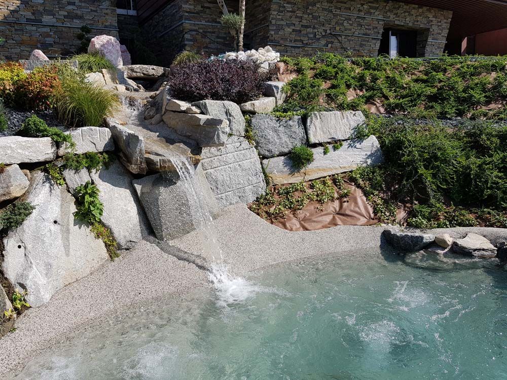Una cascata in un giardino roccioso si riversa in una limpida piscina turchese, con terrazze di pietra sovrapposte e vegetazione lussureggiante.