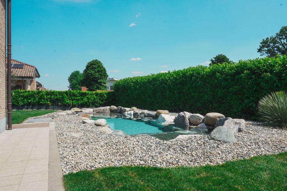 Laghetto in giardino con acqua turchese, rocce bianche e un'alta siepe verde sotto un cielo azzurro brillante