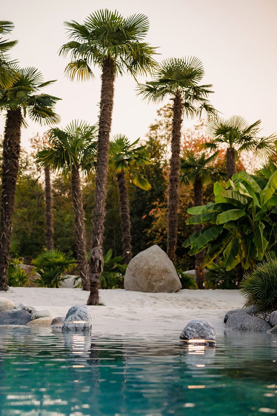 Piscina in stile resort tropicale con acqua turchese, palme e una grande formazione rocciosa al tramonto