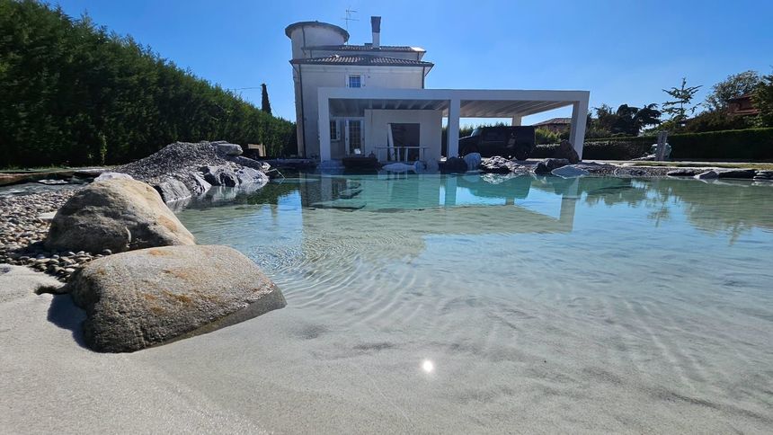 Una piscina poco profonda e limpida accanto a un moderno edificio bianco, con grandi rocce e alberi sotto un cielo azzurro brillante.