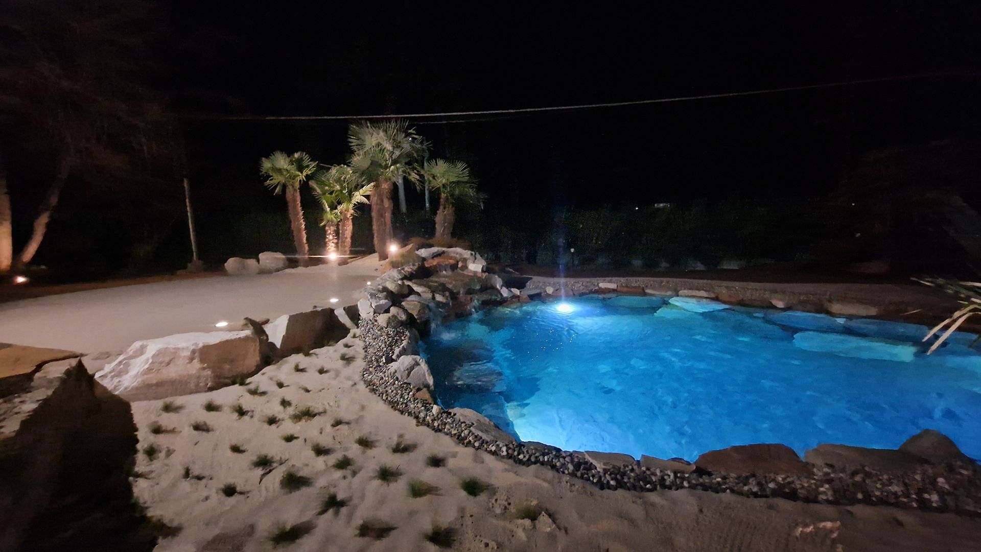 Piscina notturna in giardino con acqua blu brillante, bordo in pietra e zona salotto con patio illuminato.