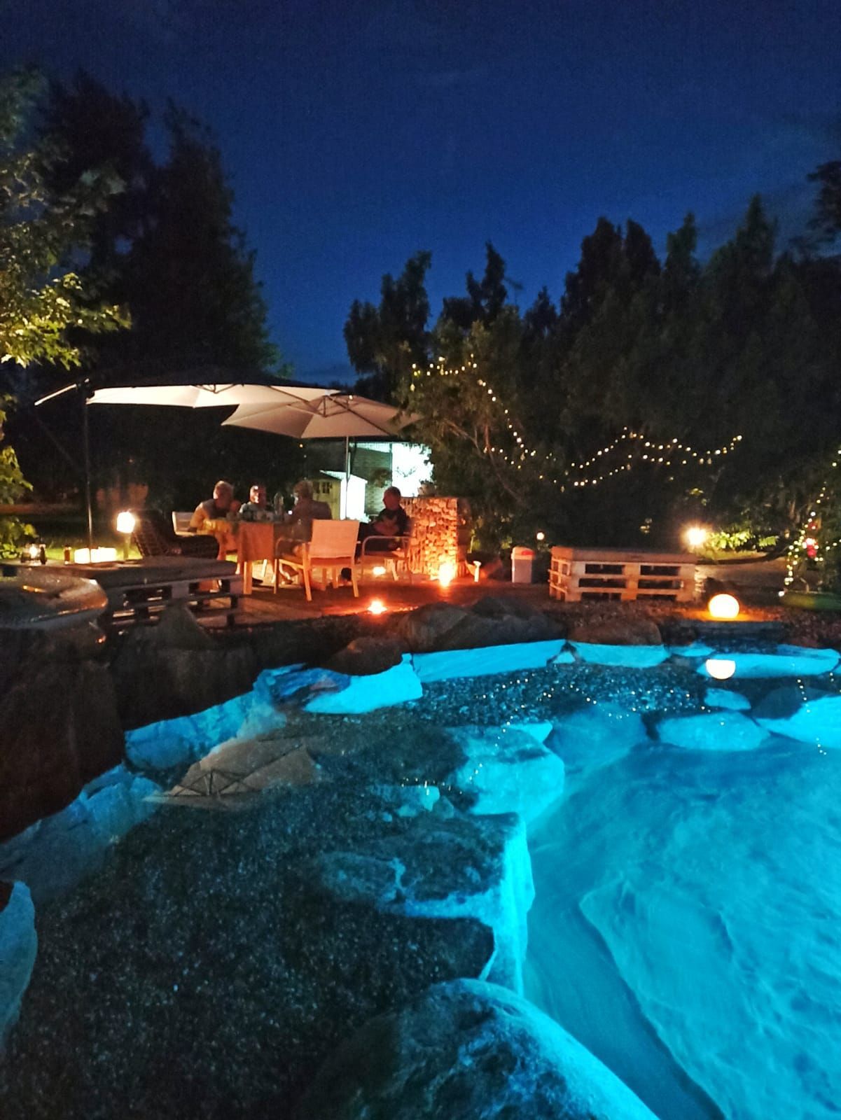 La piscina nel cortile sul retro si illumina di blu di notte, con luci da patio, alberi e sedie a sdraio intorno.